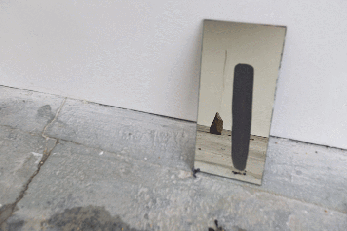 a small mirror is propped against the wall of a gallery. There is a stripe of grey paint on it and the reflection of a rock in it.