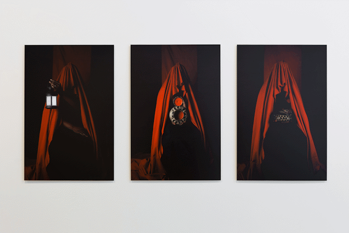three darkly lit portrait photographs of a figure swathed in red fabric, a black persons hands emerge from the fabric holding a lamp, a sculpture, a box
