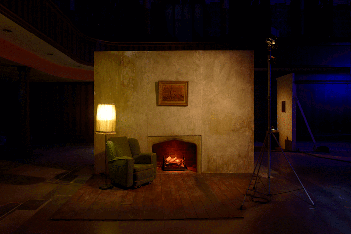 Wooden board floor, with a green chair sofa and a standing lamp switched on besides it. There is a fireplace and a painting above it. It is all illuminated by on big spotlight with a warm yellow light. We can see other movie sets in the background.This is part of an art installation in Film City, Glasgow International 2018.