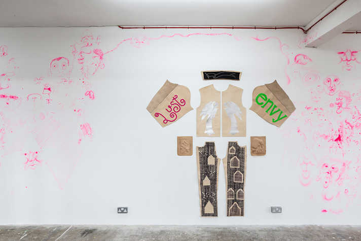 A pattern of a shirt and trousers is laid flat on a wall, decorated with art including the words 'lust' and 'envy' . On the white wall around the piece are drawings in pink.