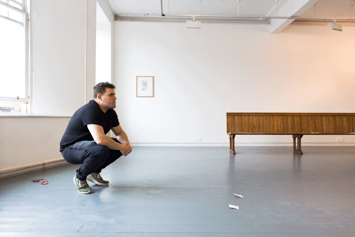 a caucasian person with short brown hair and stubble crouches in a gallery.
