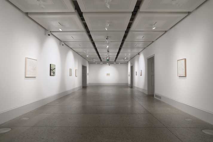9 paintings and drawings hang at a distance in a room of a white walled gallery