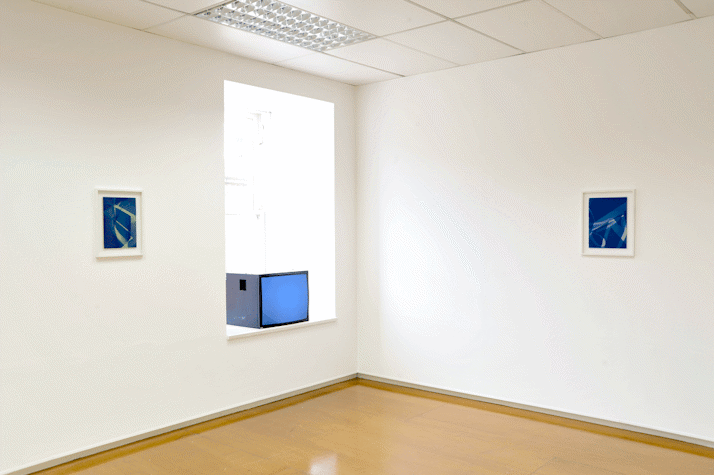 gallery with two framed cyanotypes on the left and back walls. in-between the two there is a window ledge with a cube monitor with a blue screen.