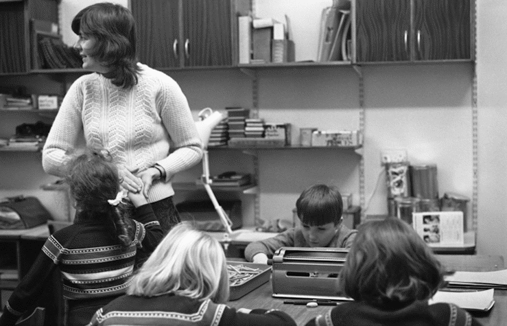 A black and white image of a room filled with a group of children sitting around a table. A woman stands next to the table holding one of the children's hands.