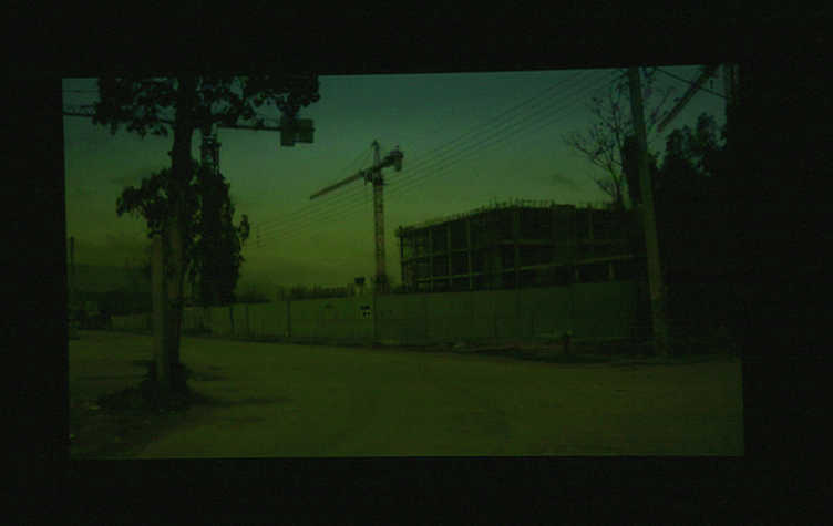 dim green and blue image of a road with a building site behind it. there is a silhouette of a crane.