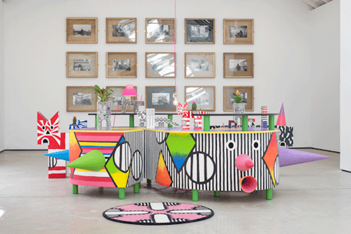 A white-walled gallery with a grid of black and white photographs on the wall behind a some multi-coloured sculptural objects in the foreground
