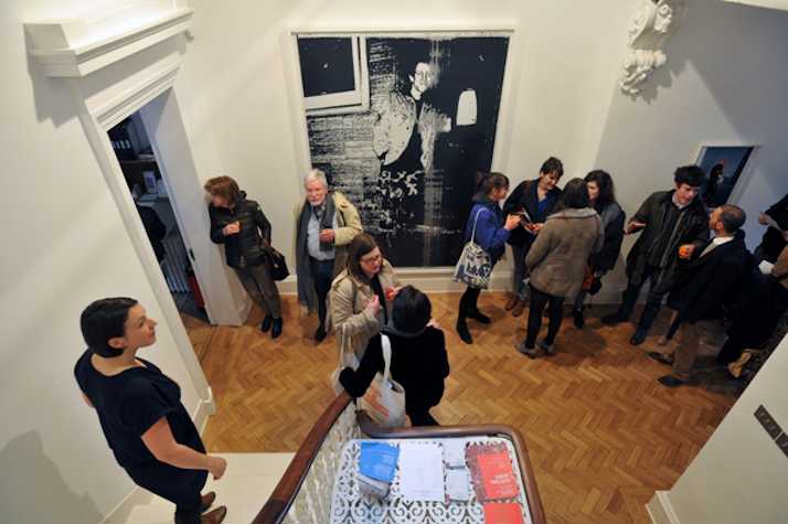 a large black and white photograph of a man is hung in the centre of a wall. onlookers surround it, speaking in small groups