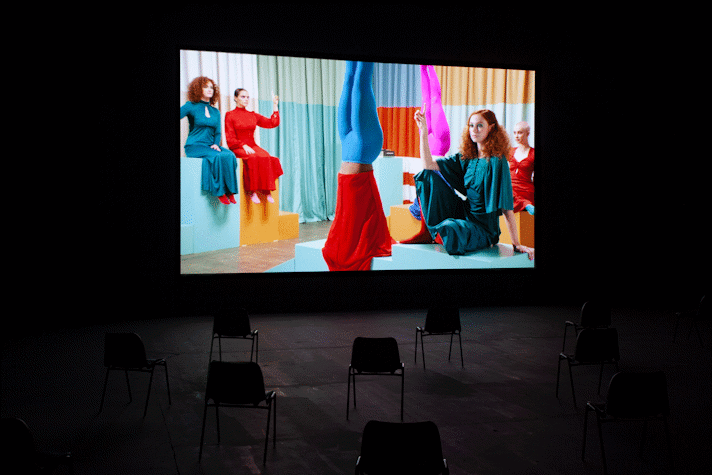 image of film being screened in a dark room. There are 4 actors in seated positions and two doing handstands. They are wearing very brightly coloured clothing.