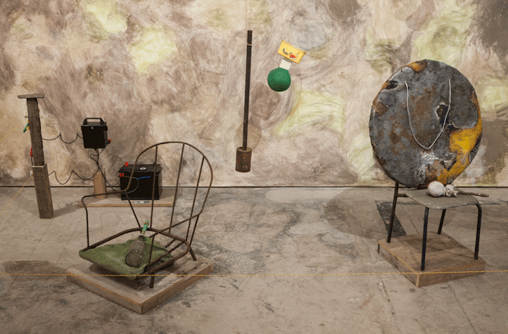 By the wall of a room, the wall is covered with paper and painted on. There are multiple sculptures placed in the room. There are some chairs, bottles, and a sculpture hanging from the ceiling, among others.