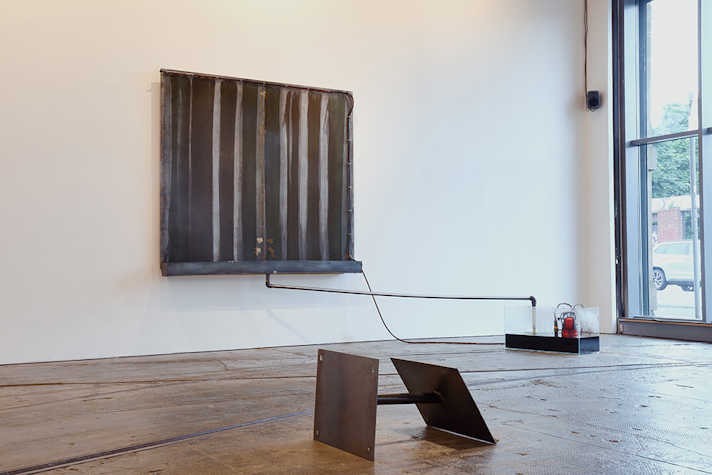 A metal sheet is on a white wall. From the bottom of the sheet is a pipe, which feeds into a clear box sitting on the group.