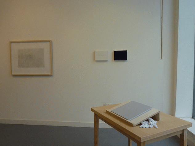 foreground shows a wooden table with an artists book resting atop with a pair of white cotton gloves. behind on the wall there are two small canvases with a larger framed work to the left.