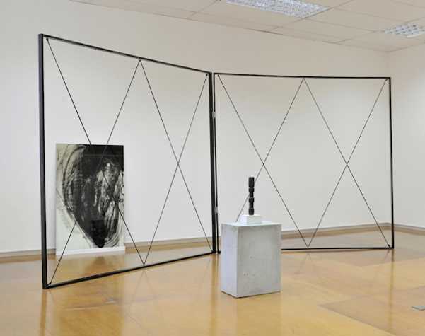 sculptural installation in a gallery space of a criss-crossing structure in a frame. in front there is a plinth with a small black sculpture. behind there is an expressive painting leaning against the wall.