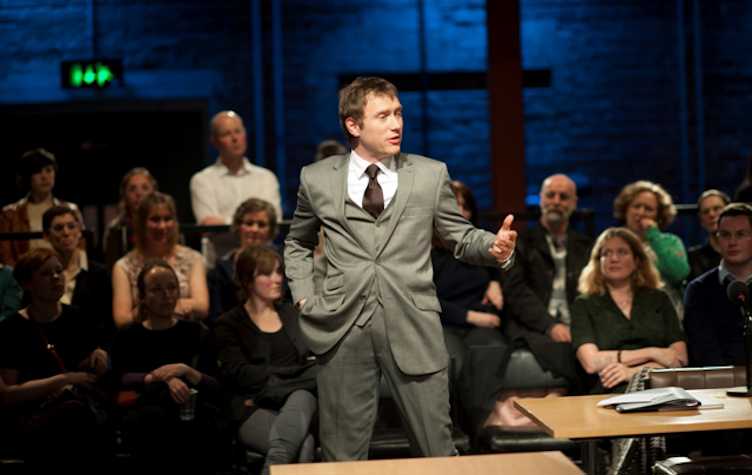 man wearing a grey three piece suit standing in front of an audience