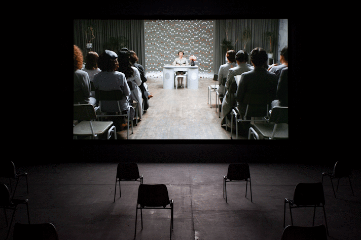 An image of a film being screened in a dark room. The screen shows a person at a desk with an audience in front of them. There is a curtain of eggs suspended in the air behind them.