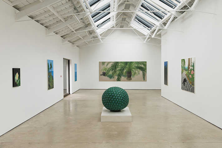 An installation view of a exhibition featuring artworks on white walls, in a well-lit space. In the centre of the room is a large green sphere.