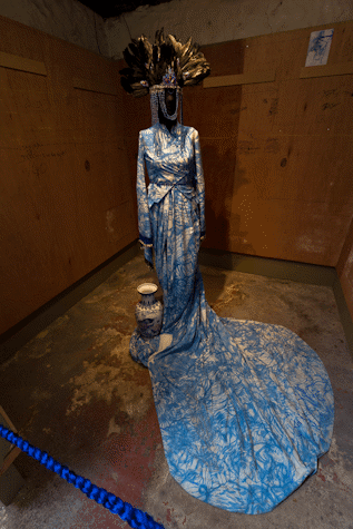 A manequin with an intricate dress made from fabric with an abstract indigo painted pattern. The dress has a long train that is draped around the front of the manequin, upon which a vase sits. The manequin wears a headdress of large black feathers with strands of beads draped around the face.
