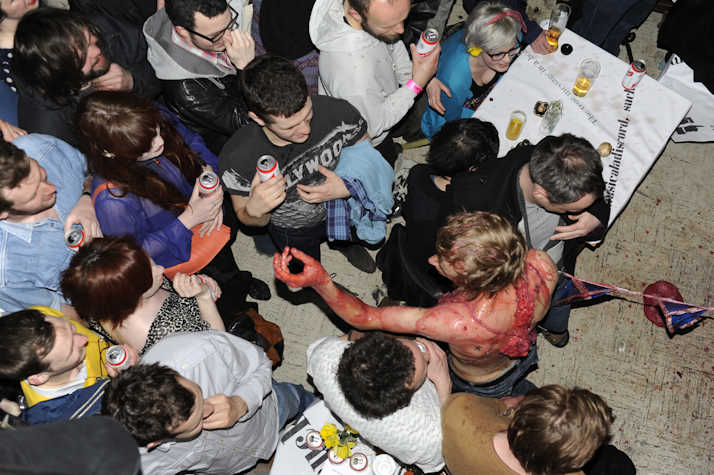 Crowd gathered around a performer with red body paint and stage effects, holding drinks. View from above, with people interacting and watching.