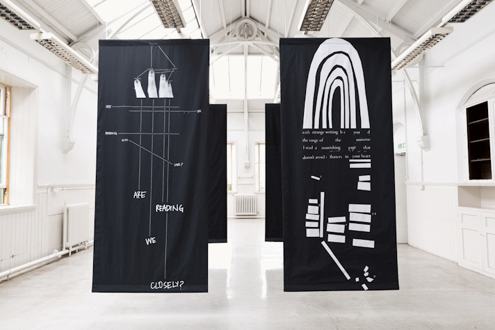 four portrait orientation large black rectangles of fabric hang from the rafters of a large school hall-esque room. The front two have white printed text, diagrams and shapes on.