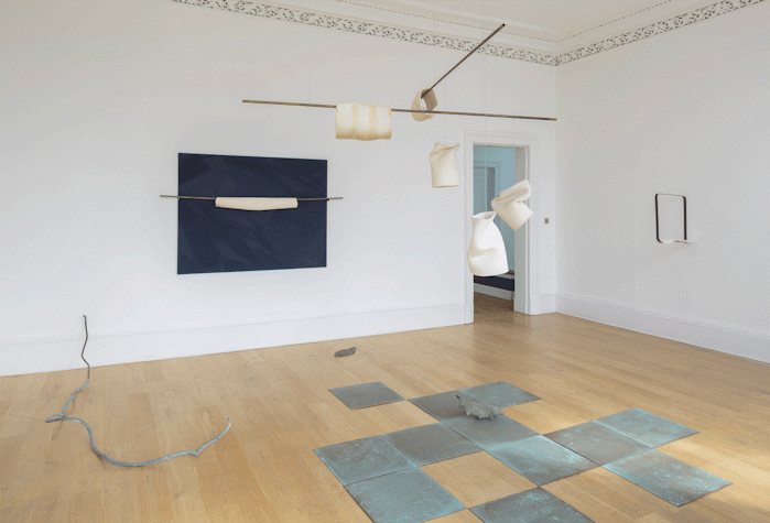 Room with wooden floors and a grey ceramic figure in the ground as well as ceramic tiles. Several white figures hanging from the ceiling and a dark blue artwork in the back wall.