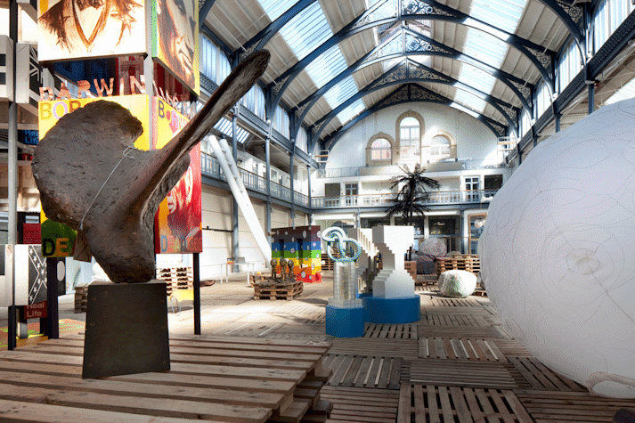 In a large hall, with a glass ceiling, there are multiple sculptures. There is a blimp like baloon on one side, and a large hip bone like structure on the other. There are some very colourful and bright sculptures, while some have the shape of an hourglass.