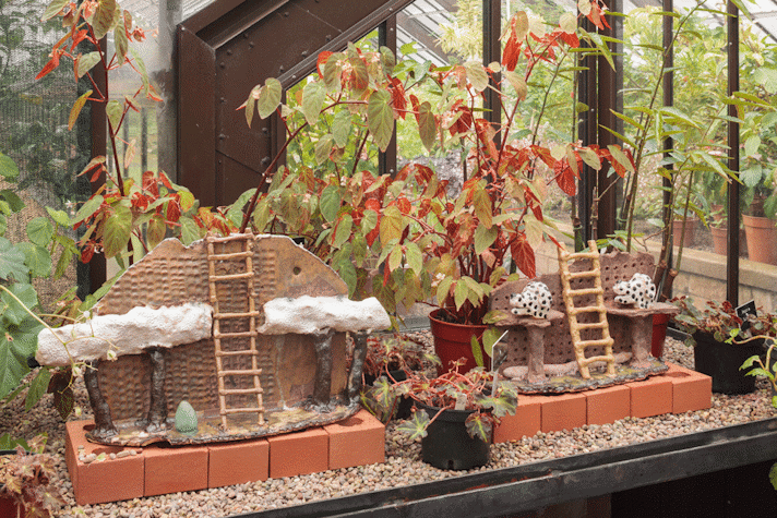 In a greenhouse, between plants, two small displays of ceramic trees with a ladder in between, in front of a base of bricks.
