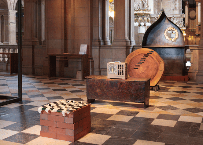 A chess display on top of a table made of bricks, behind it, we can see a metalic cream colored box on top of a wooden storage box. Behind that, a big cut tree rings with black labels on it. Behind, a metal conical structure with the see-through circle surrounded by white flowers.