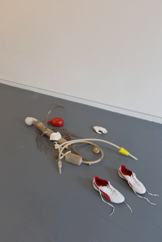 sculptural assemblage on the floor of a gallery. There are trainers, sticks, plastic tubing, a hand-like object.