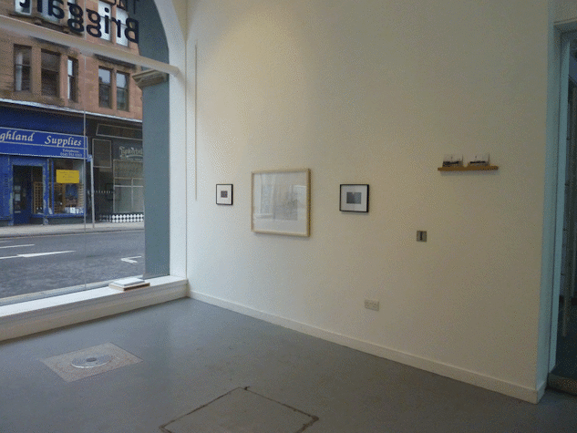 three framed works on a gallery wall and two small postcards on a wooden floating shelf. to the left is a large glass window showing the street outside.