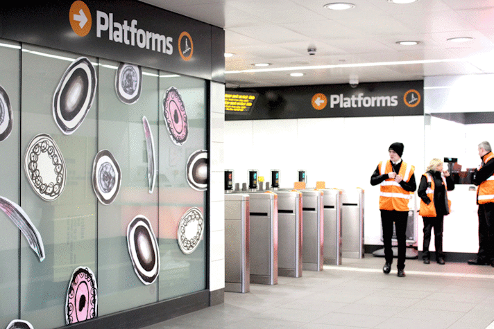 The subway entrance of Ibrox has circular drawn figures in the wall, in black, white and pink colours. Three subway station workers in their uniforms and orange vests are standing around the ticket entrance.