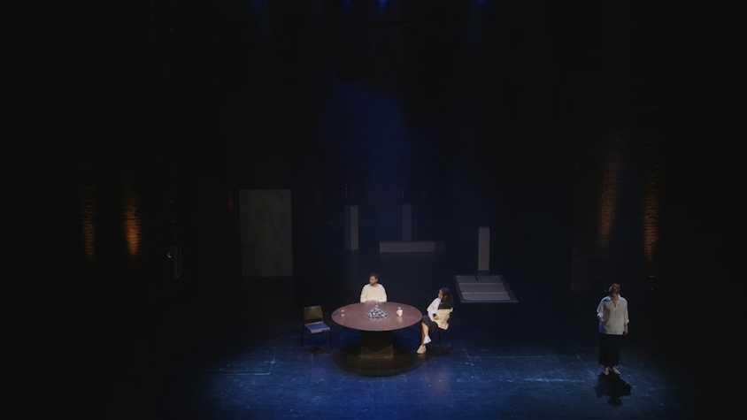 wide shot of tramway 1 stage. two people are sat at a round table in the centre whilst another stands off to the side