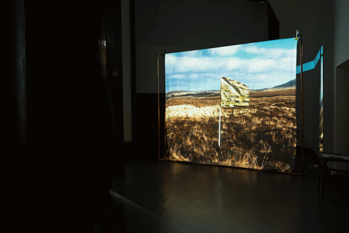 In a dark room a film is projected onto a screen. The project image shows a flag made from a gold emergency blanket, flying in an open field with hills in the far distance and a blue sky