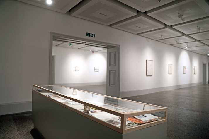 A glass vitrine sits on a large white plinth. Inside the vitrine are a collection of open sketchbooks and books. Behind it a gallery room stretches out with small scale paintings and drawings hanging on white walls.