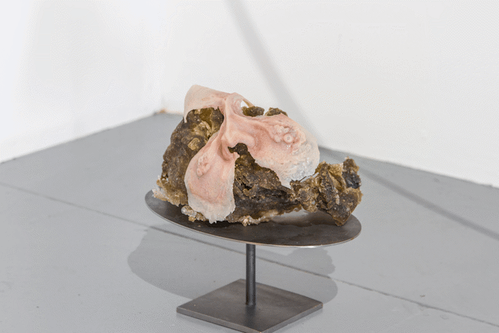 On top of a metal bronze table an irregular brown rock is mixed with a material that ressembles pink flesh.