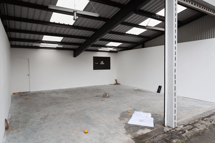 objects including a white towel and oranges are strewn across a concrete floor space. In the background a dark print is pasted to the wall