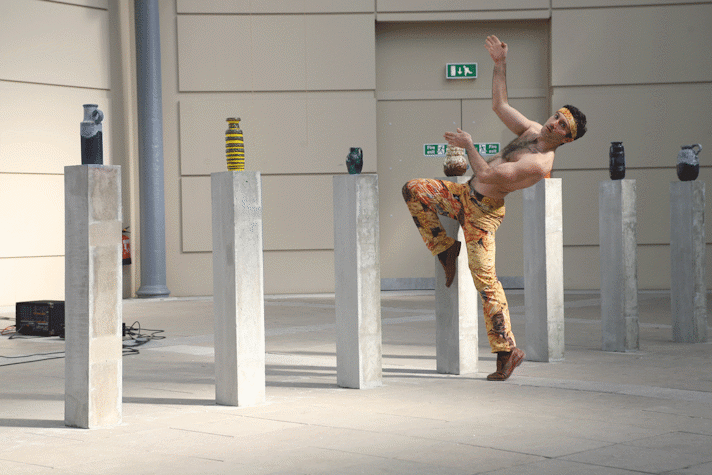 man wearing trousers covered in images of pasta captured on one leg, mid motion. behind him is a row of concrete plinths with various ceramic jugs and vases atop.