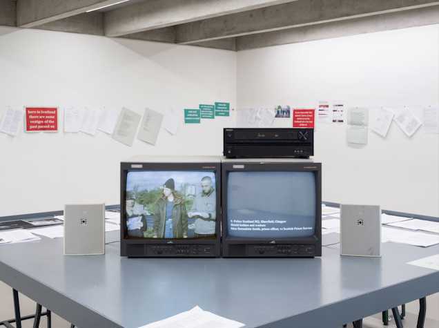 A photo of a room with a grey table with two TVs sitting on it, the left screen has three people standing outside and the right screen is black with white text reading 'Police Scotland HQ, Shawfield, Glasgow.' In the background of the image are white walls covered by a series of signs and pieces of paper.