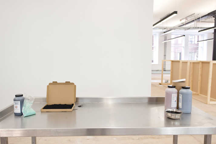 A metal table has several materials on top of it: a carton box, a small bowl, pigment and bottles.