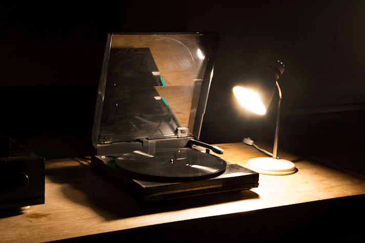 On a table, with a table lamp turned on and shining light on a vinyl record player. There is a vinyl record inserted in the player.