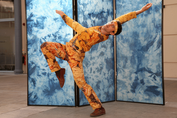 man stands on one leg, leaning back, moving expressively wearing a suit patterned with images of pasta in front of a trifold screen with blue marble patterning.