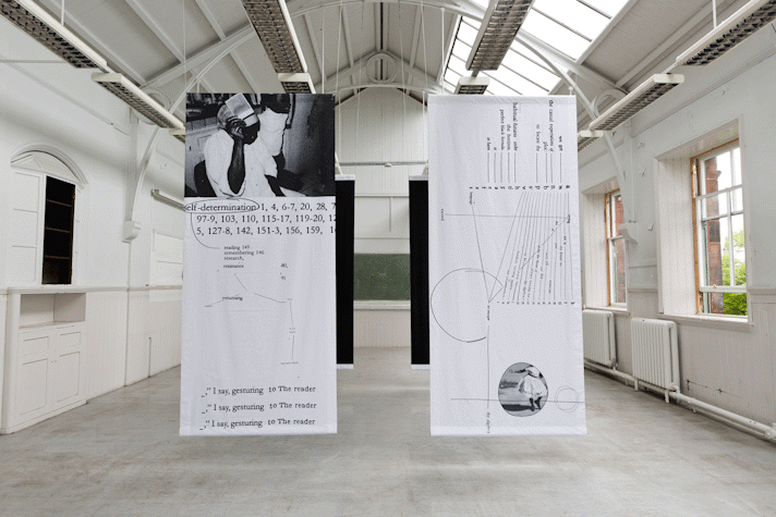 four portrait orientation large rectangles of fabric hang from the rafters of a large school hall-esque room. The front two hangings have black text, numbers and diagrams on and a black and white photograph of a black person with white shirt and holding a postcard in their hand