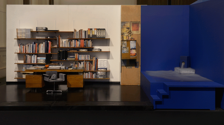 An office with a white wall has a shelves stacked with books. To the right is a small stage all painted dark blue with fog coming out of it as part of an art installation in the Gallery of Modern Art, Glasgow International 2018.