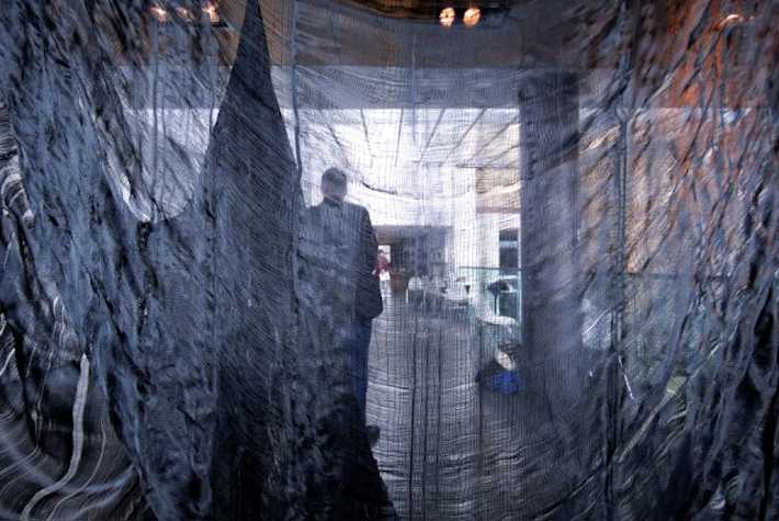 image taken through blue crinkly sheer fabric. in the background you can make out the silhouette of a person and some chairs and tables in the far back.
