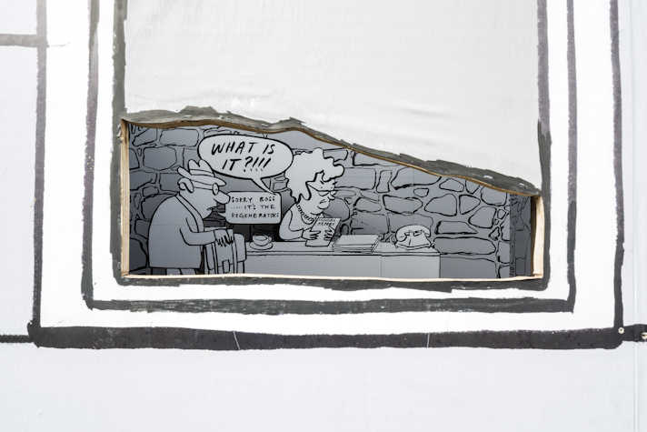 Through the painted window frame we see another drawn black and white mural: an office with a stone wall in the background. The boss has curly short haired and glasses and looks upset. A speech bubble emerges from them saying "WHAT IS IT?!!!". To the left there is a hunched man with glasses and a hat holding a briefcase. His speech bubble reads "Sorry boss..... it's the regenerators".