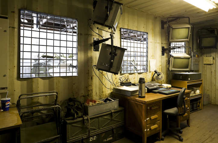 Interior of a surveillance room in a shipping container, featuring a desk with monitors, a chair, and equipment. Gridded windows reveal the adjacent exhibit space. Stacked crates and old security screens are mounted on the walls.