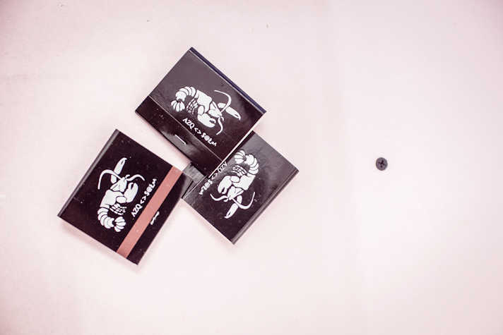 On a blank background, there are three sachets with a scorpion printed on them. They carry the title AZQ < > $@Ł · ^ under the image of the scorpion. The middle sachet is staple. There is a nail screwed on the background next to the sachets.