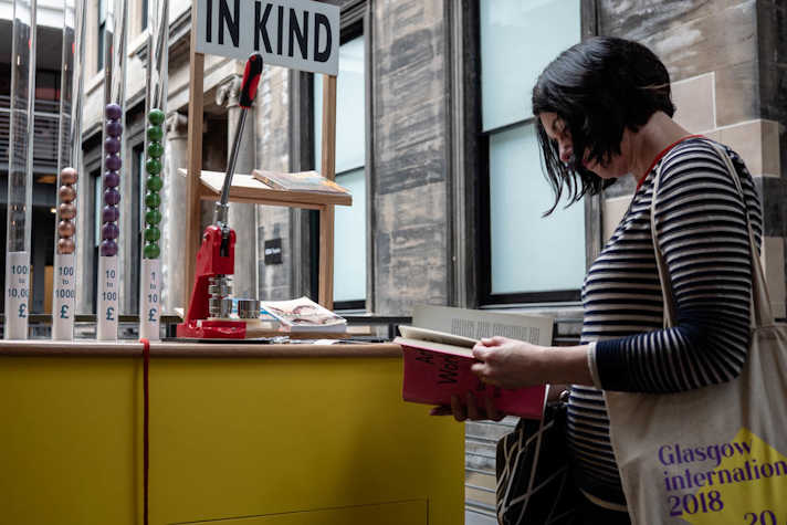 In the CCA a white woman with short black hair and a stripped black and white sweater is standing in front of the IN KIND stand, reading a pink book that says "Art Workers". She has a tote bag that reads "Glasgow International 2018".