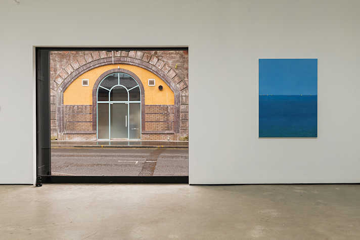 An artwork featuring blue colours is located next to a large window looking onto a doorway.