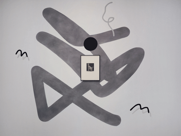 A white wall with some abstract marks applied directly, a framed image in the middle, and two small three-dimensional sculptural works in front of the wall