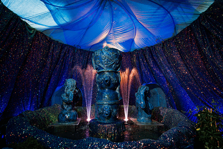 A blue water fountain featuring ceramic nude figures is underneath a domed blue ceiling with walls decorated by sparkly blue fabric.