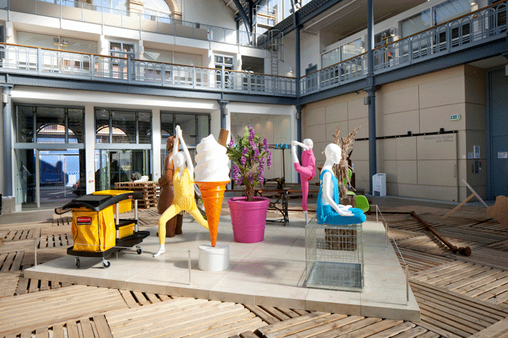 In the corner of a large hall with a glass ceiling, there are multiple sculptures standing on a marble platform on a temporary wooden floor. There is a large ice cream sculpture, next to a three people doing different yoga poses.